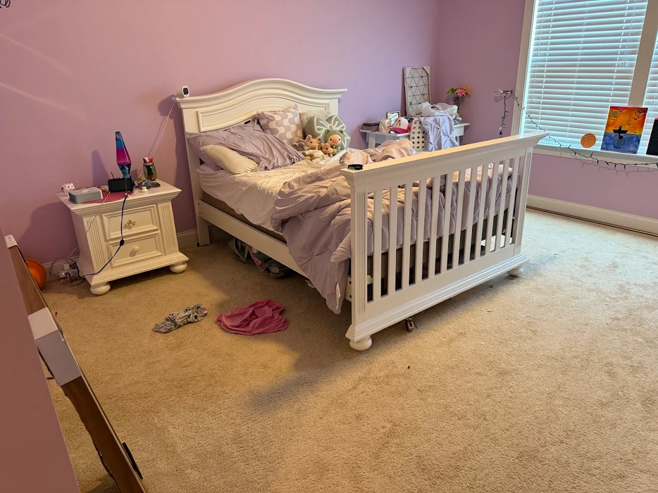 A child's lilac bedroom before cleaning, with an unmade bed and clothes on the carpet
